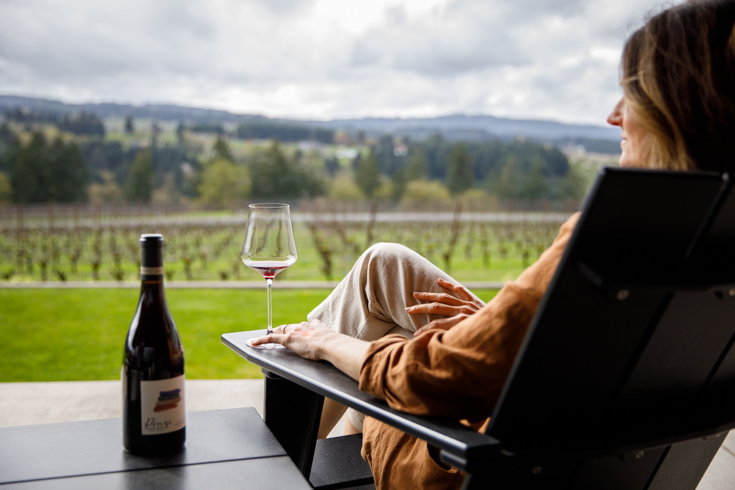 Person sitting at a table with a bottle of wine and a glass, overlooking vineyards.