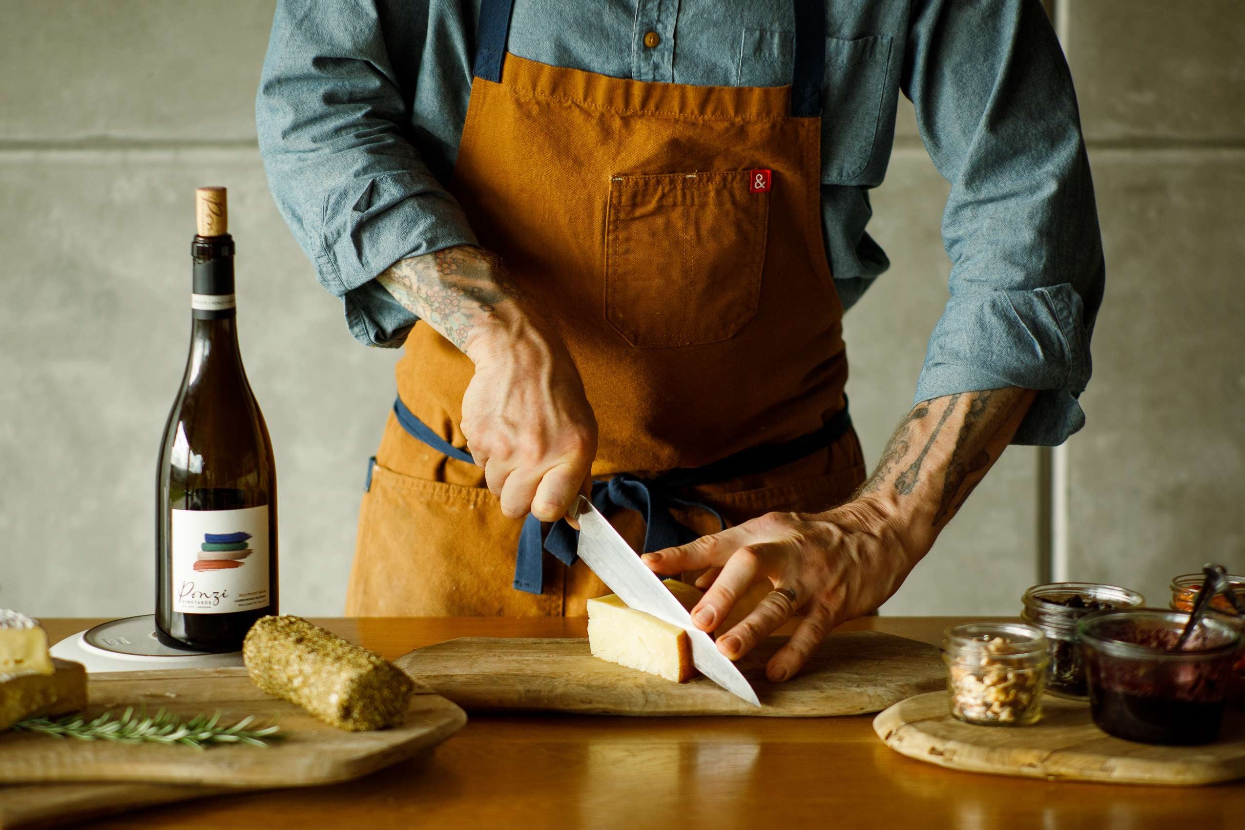 A person cutting cheese on a wooden cutting board, surrounded by various ingredients and a Ponzi Vineyards wine bottle.