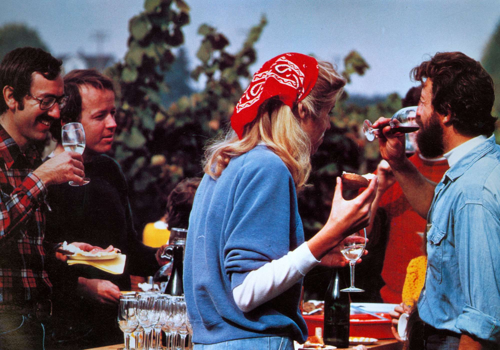 Group of people outdoors, one person in a blue sweater and red bandana is holding a wine glass.