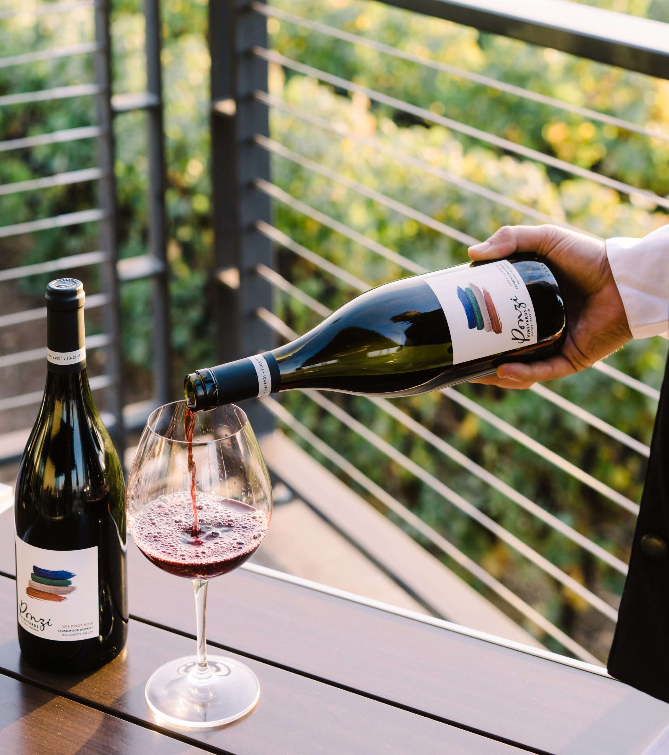 Person pouring red wine from a bottle into a glass on a balcony with greenery in the background