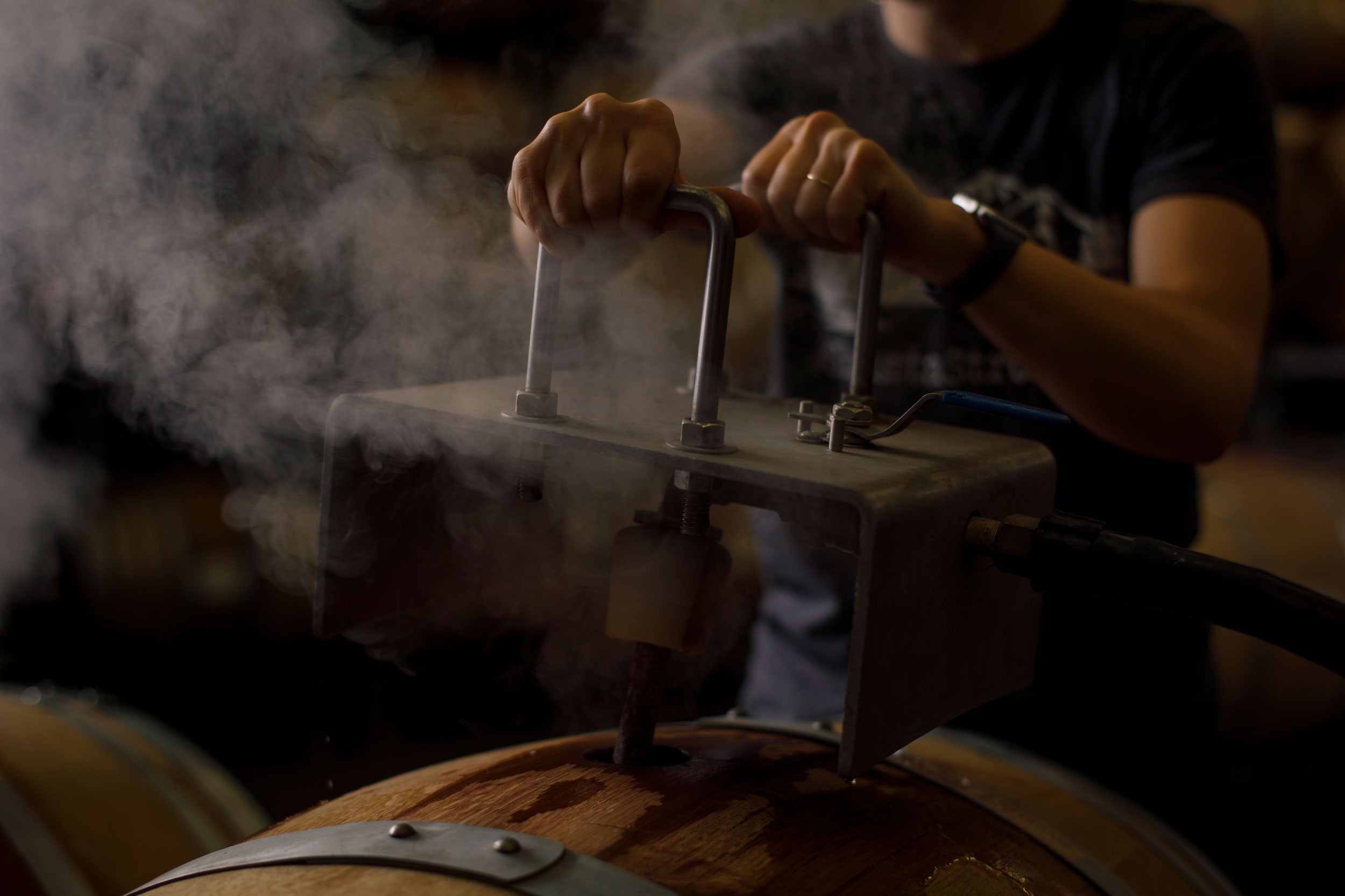 Person operating a steam press on a wooden barrel with steam rising, in a vineyard setting.